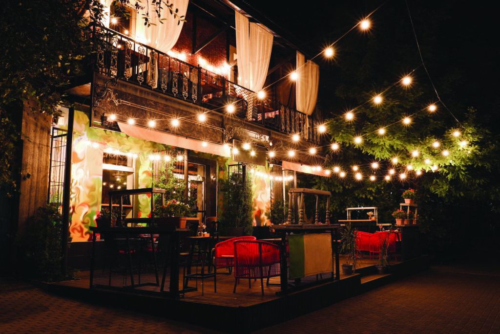 Cozy outdoor restaurant patio at night decorated with string lights, red chairs, and curtains, creating a warm, inviting atmosphere.