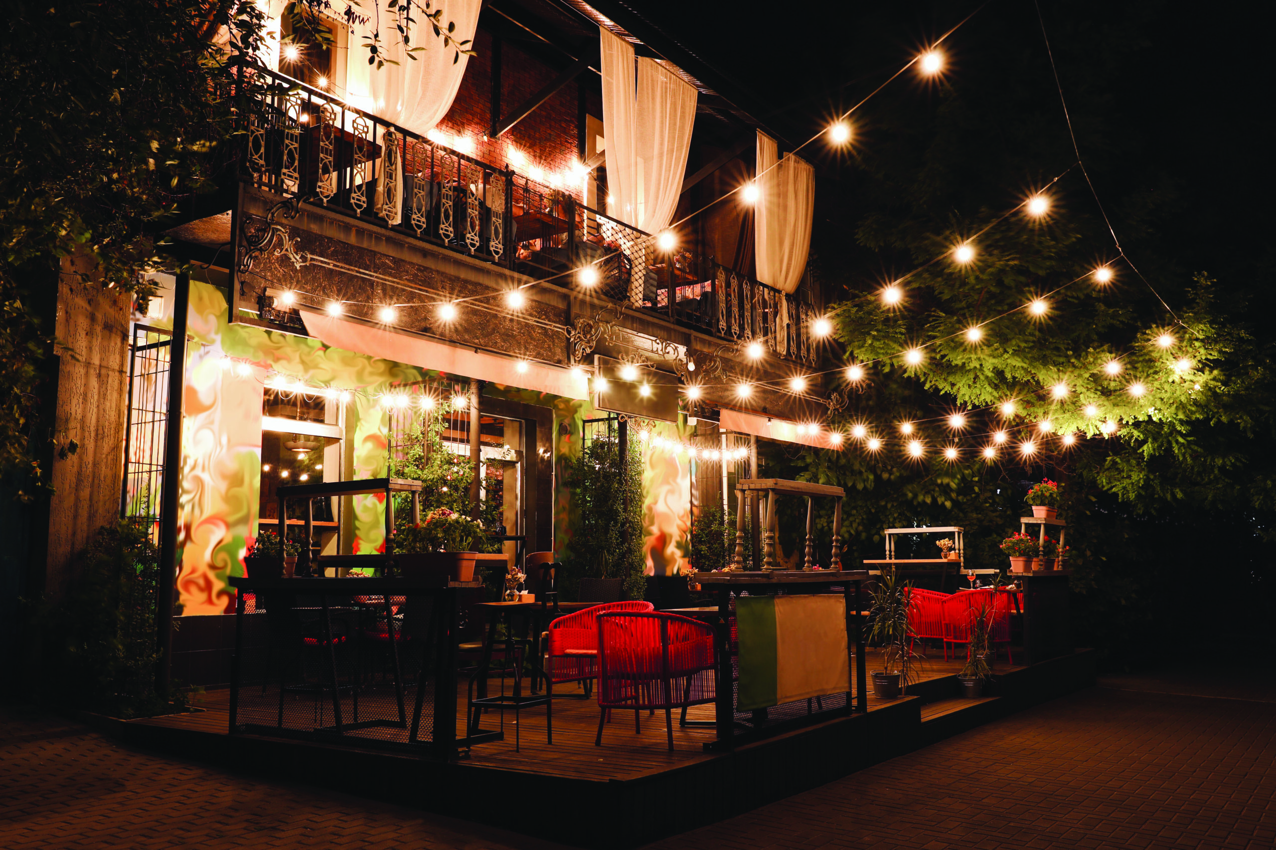 Cozy outdoor restaurant patio at night decorated with string lights, red chairs, and curtains, creating a warm, inviting atmosphere.