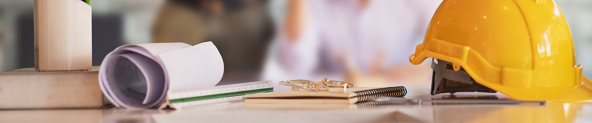 Architect’s desk with a yellow hard hat, rolled blueprints, and documents, with a person working in the background.