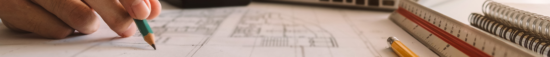 Close-up of a person sketching or marking on architectural blueprints beside a scale ruler and notebooks.