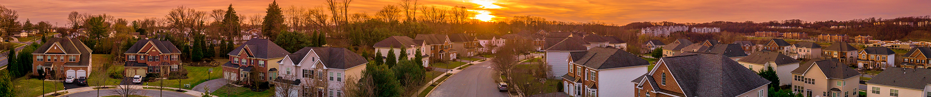 Aerial view of a suburban neighborhood at sunset, with rows of houses and tree-lined streets under a warm orange sky.