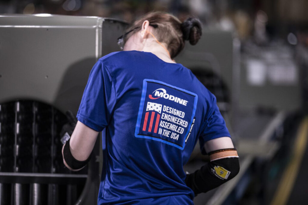 Factory worker wearing a blue “Modine – Designed, Engineered, Assembled in the USA” shirt inspecting large industrial equipment inside a manufacturing facility.