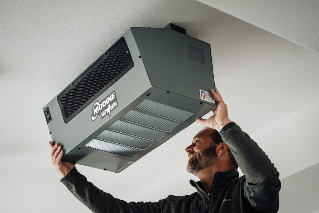 Man installing or inspecting a Modine heating unit mounted on a ceiling, smiling as he adjusts the equipment in a bright indoor space.