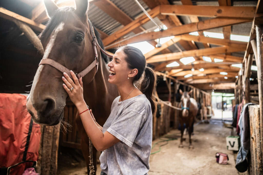 A smiling young woman pets a brown horse inside a rustic wooden stable with another horse standing in the background. Natural light filters through the slatted roof.