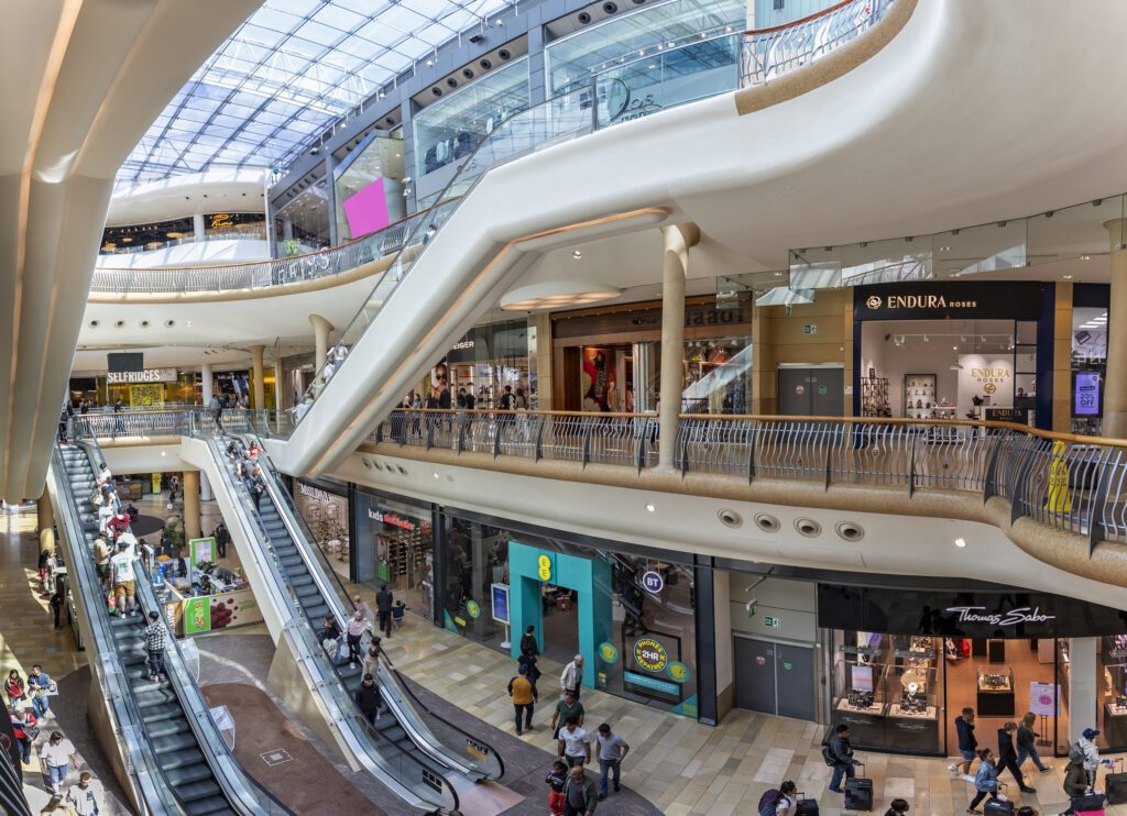 A modern shopping mall with multiple levels, escalators, glass railings, and a skylight ceiling, filled with shoppers.