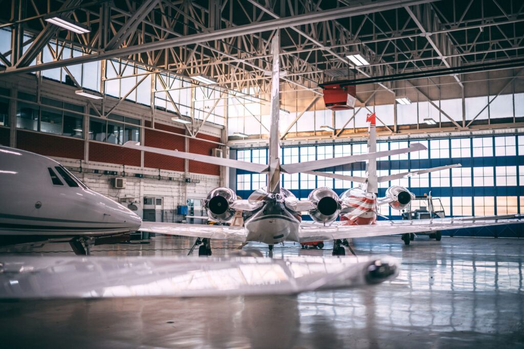 A spacious aircraft hangar with private jets parked inside, illuminated by overhead industrial lighting and natural light streaming through large windows.