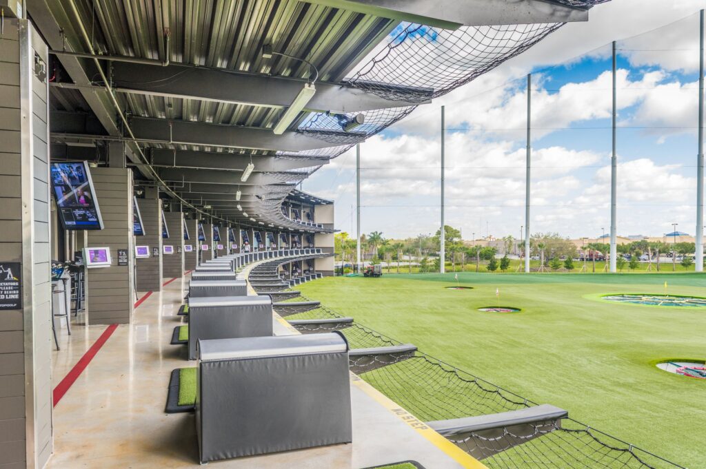 A modern outdoor golf driving range with multiple hitting bays, each equipped with seating, digital screens, and protective netting, overlooking a green field with target areas.
