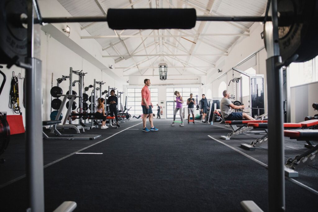 A spacious gym with high ceilings, natural light, and various workout equipment, including weight benches, squat racks, and rowing machines, with people engaged in fitness activities.