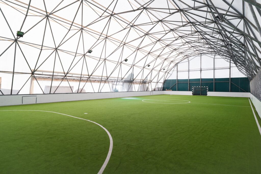 Indoor sports facility with artificial turf and a curved geodesic dome roof structure allowing natural light.