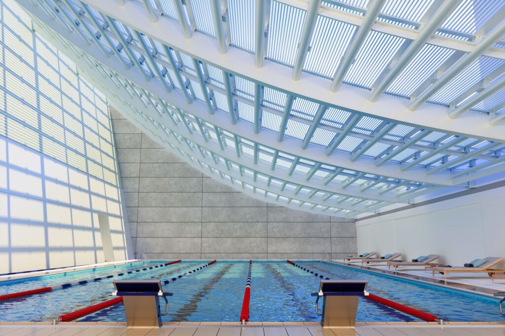 An indoor swimming pool with marked lanes, sleek modern architecture, and abundant natural light streaming through the curved ceiling panels.