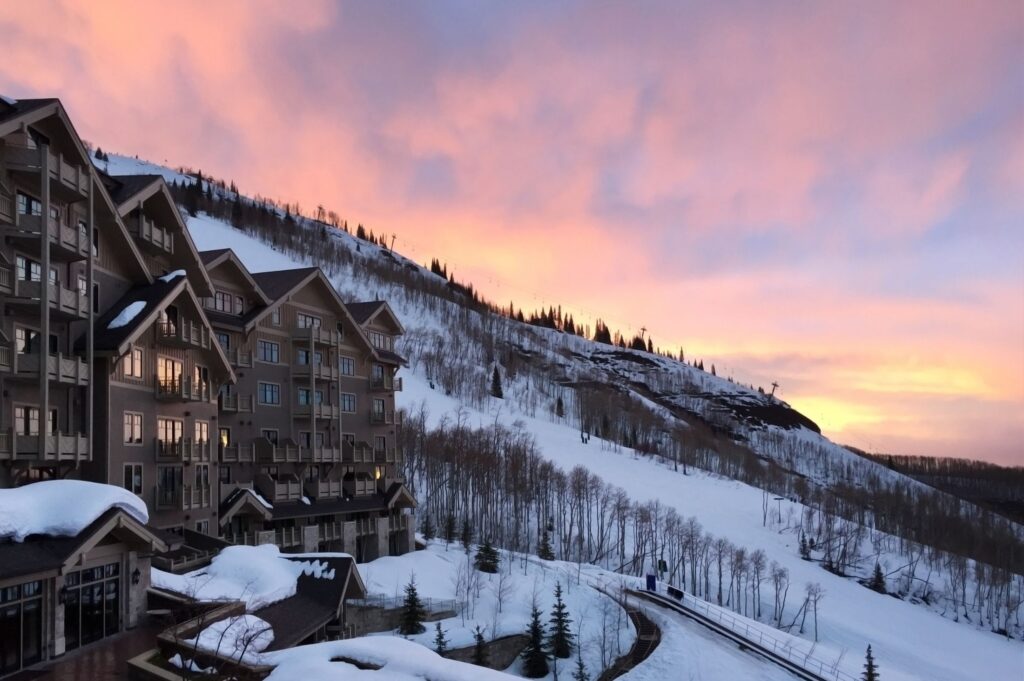 A snow-covered ski resort nestled against a mountain slope at sunset, with warm lights glowing from chalet-style buildings.