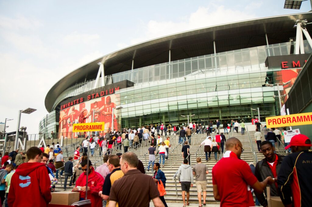 Fans in team jerseys and casual attire ascend the steps of a modern stadium with a glass facade, preparing for a sporting event.