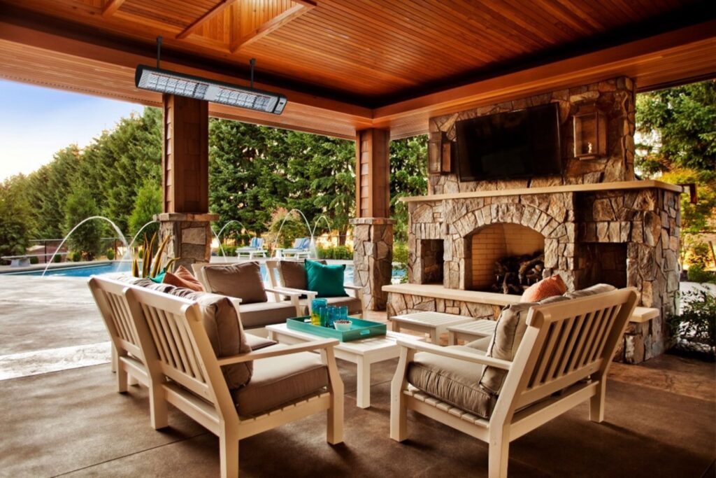 A cozy outdoor patio with cushioned white wooden furniture, a stone fireplace with a mounted TV, and a ceiling-mounted infrared heater, overlooking a landscaped pool area with fountains.