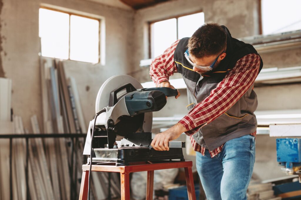 A man in safety goggles and a plaid shirt using a miter saw in a workshop with metal and wooden materials in the background.