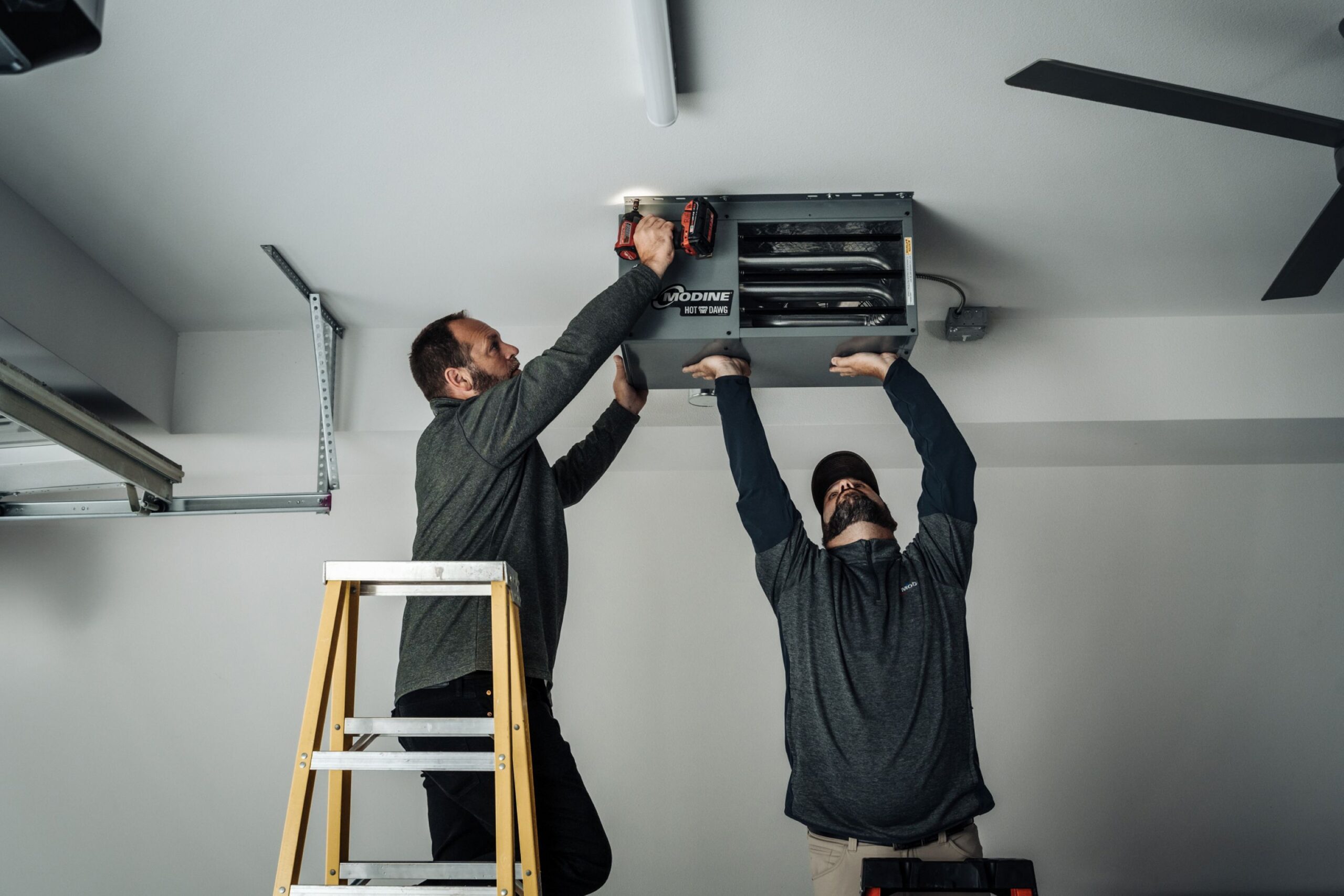 Two men install a Modine Hot Dawg heater on a ceiling; one stands on a ladder using a power drill while the other supports the unit from below.