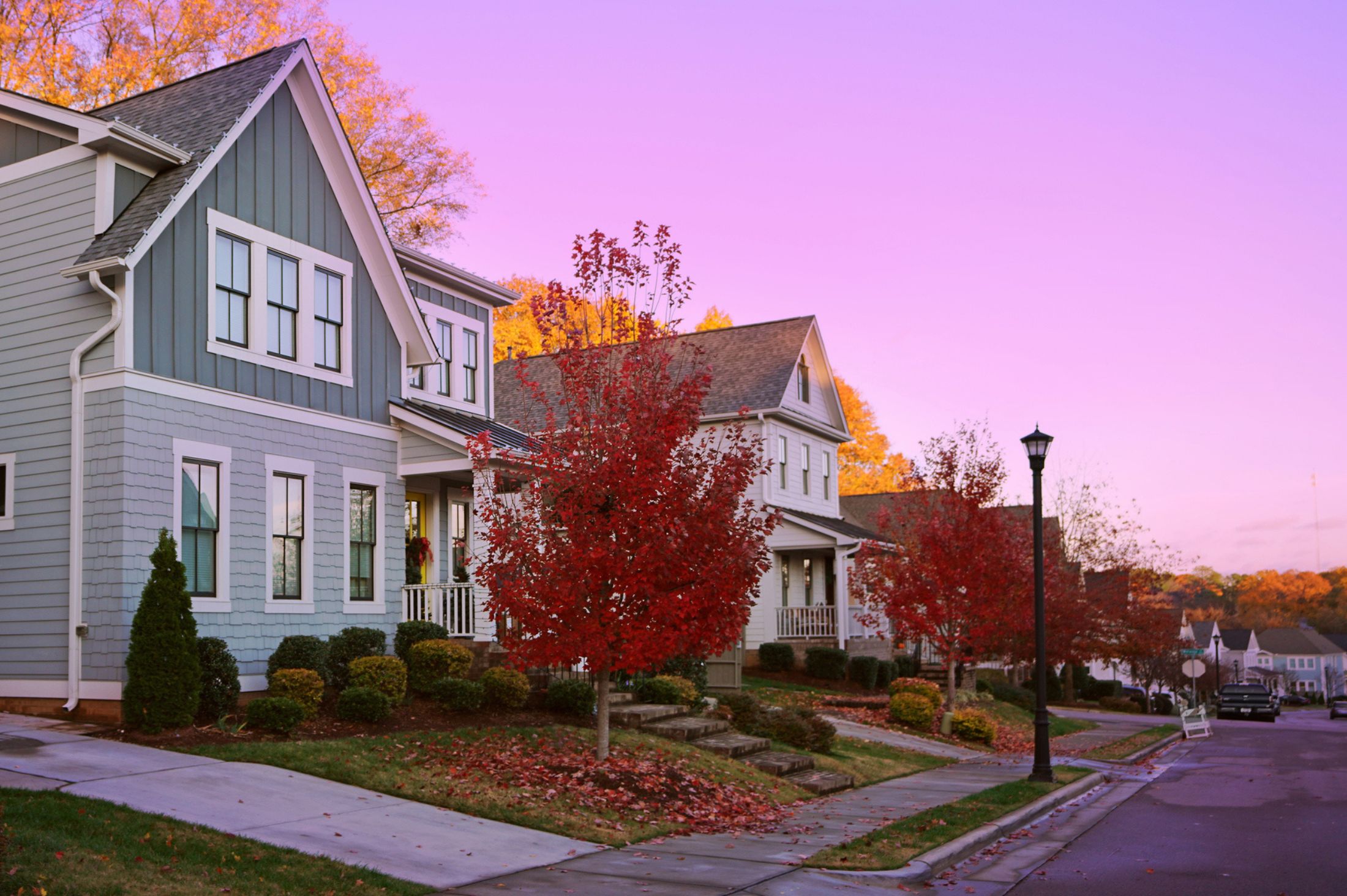 Row of modern suburban houses with gray siding and front porches, lined with red autumn trees along a quiet residential street at sunset.