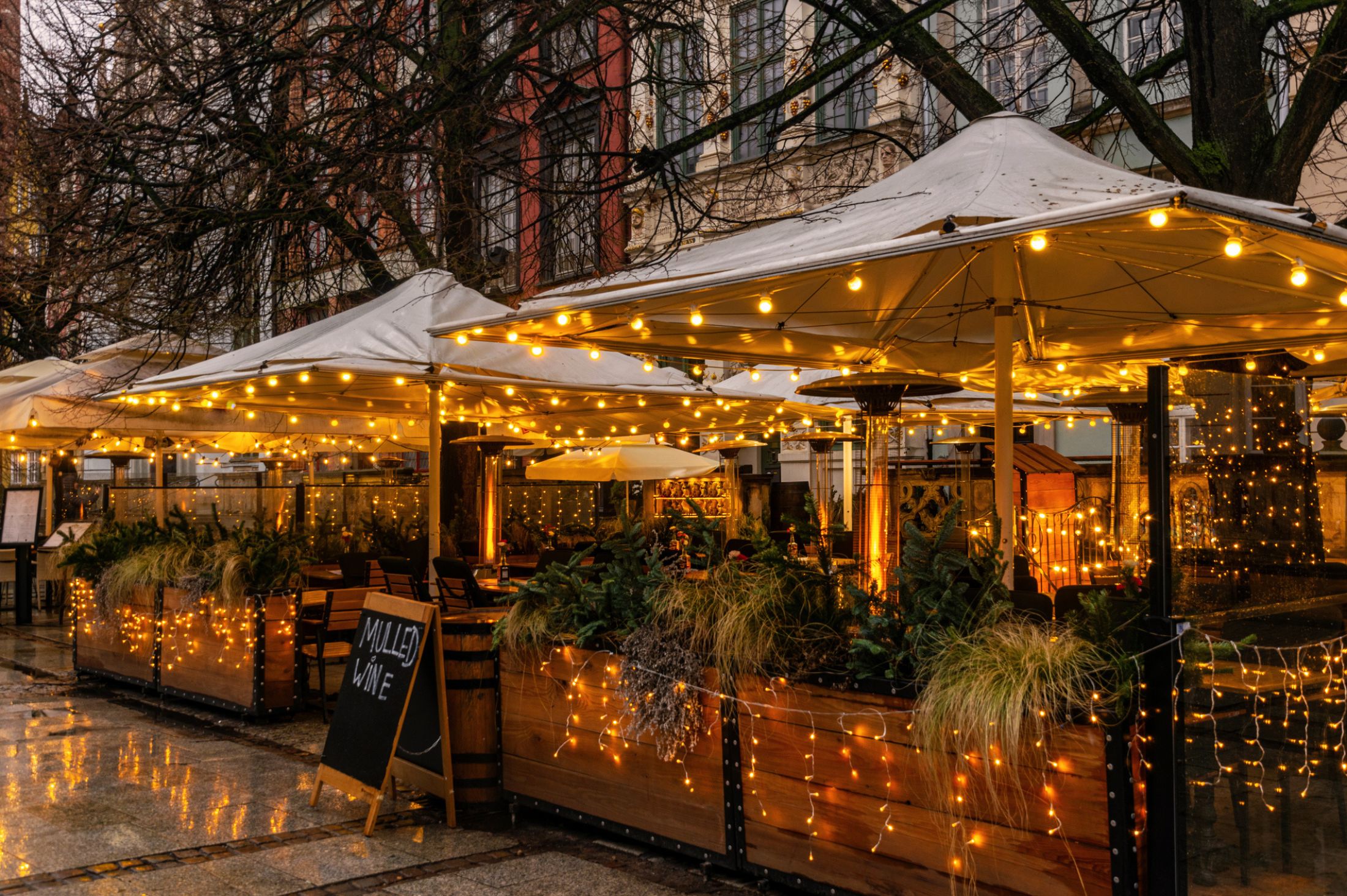 Outdoor restaurant patio with white umbrellas, string lights, and planters creating a cozy atmosphere on a rainy evening.