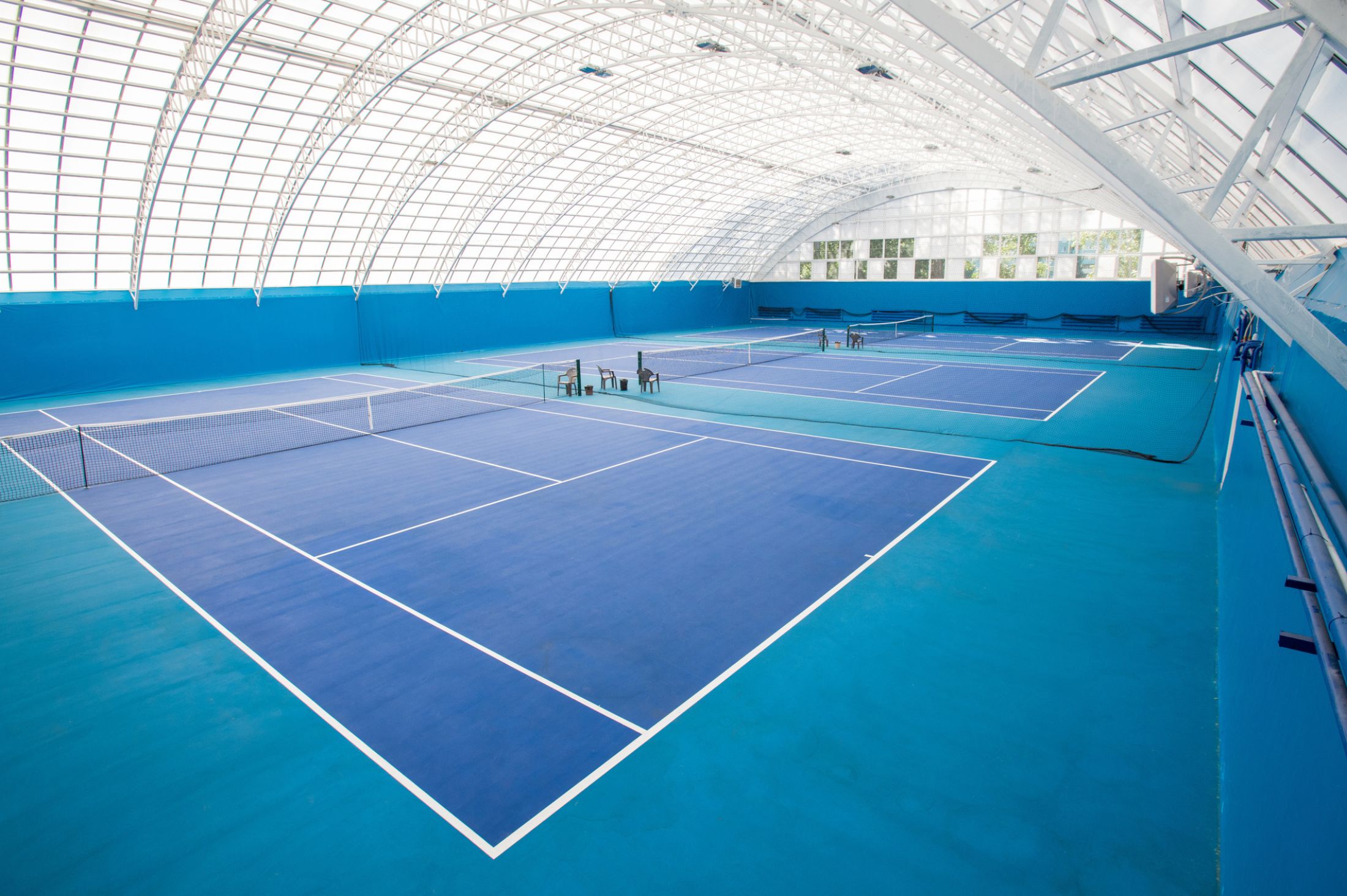 Indoor tennis facility with bright blue courts, white lines, and a high arched ceiling made of translucent panels allowing natural light.