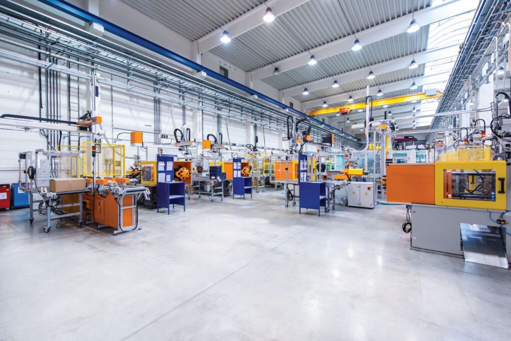 Bright industrial facility with multiple yellow and blue manufacturing machines arranged in rows beneath a high ceiling with skylights and overhead pipes.