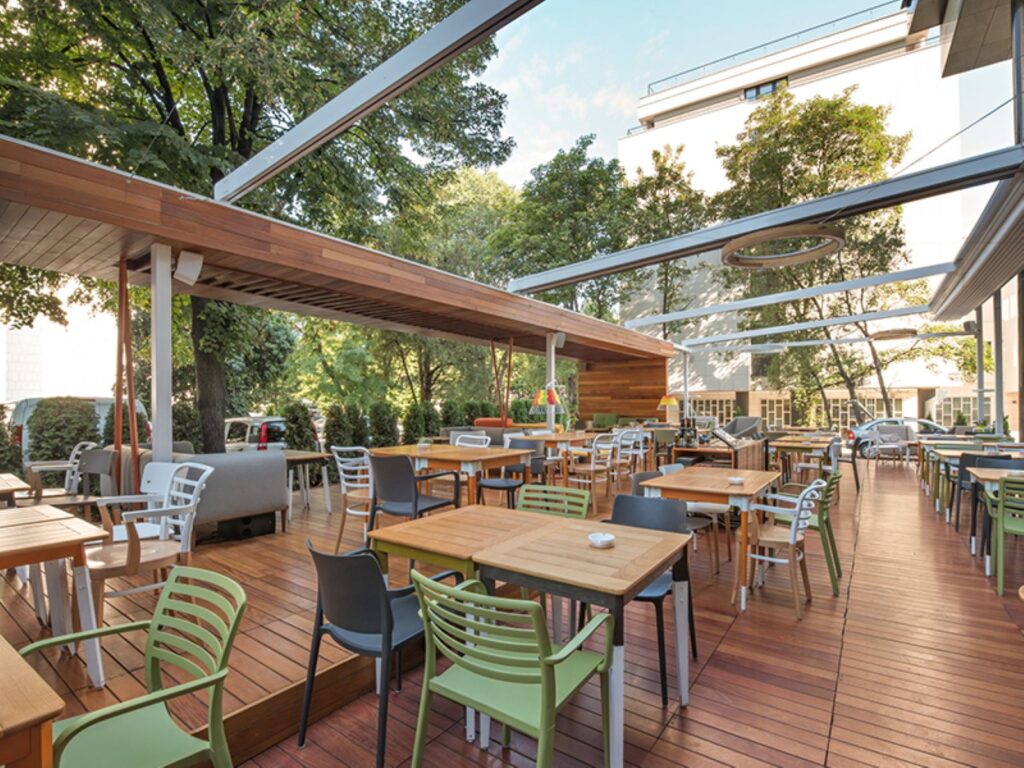Outdoor restaurant patio with wooden decking, modern tables and chairs, and partial overhead beams surrounded by trees and greenery in daylight.