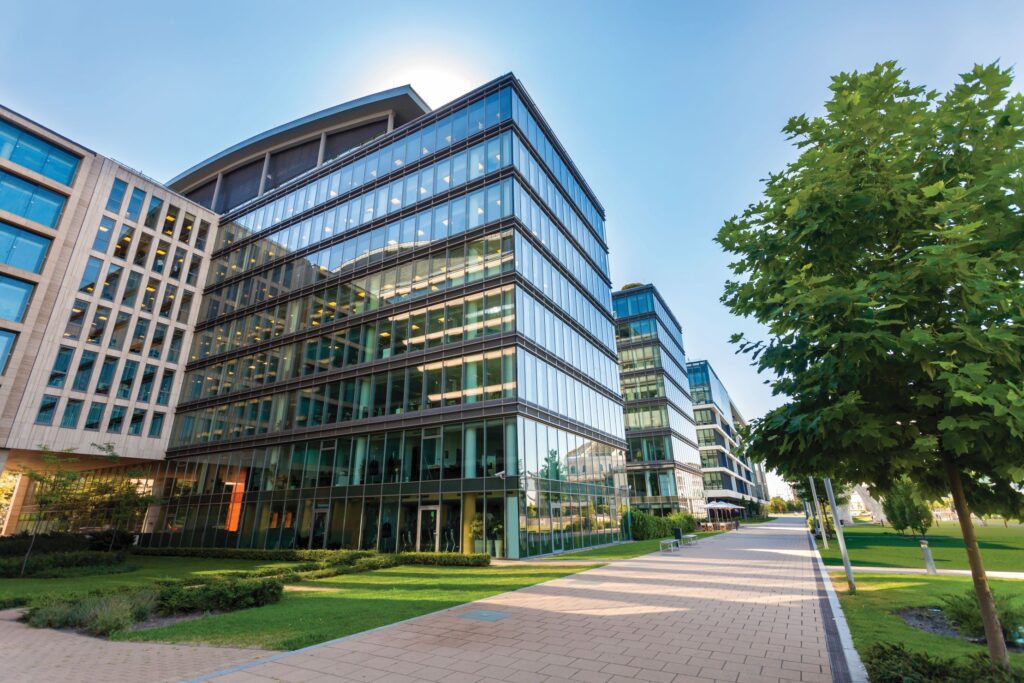 Modern glass office buildings with landscaped walkways and trees under a clear blue sky.