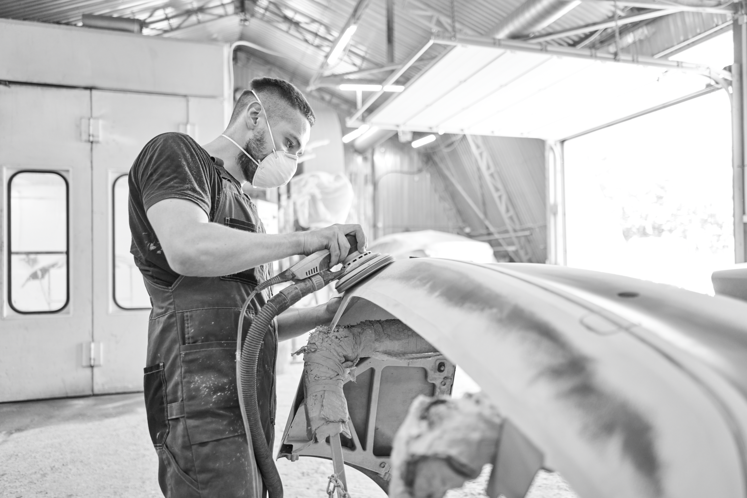 A worker in an automotive repair shop sanding a car part while wearing a protective mask, with a well-lit industrial workspace in the background.
