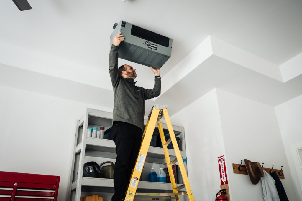 contractor installing a ceiling-mounted amp dawg electric unit heater, yellow ladder, wood garage shelving, red tool chest, a fire extinguisher and clothing hooks on the wall.